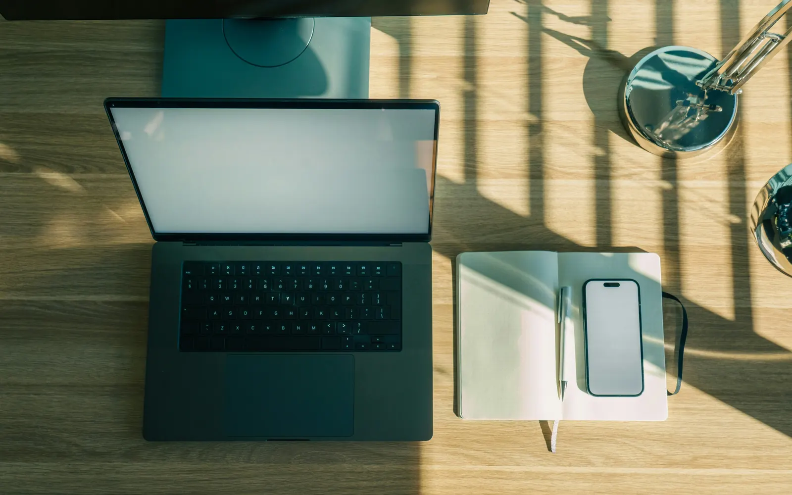An overhead view of a laptop, open notebook, and phone on a wooden desk in warm afternoon light
