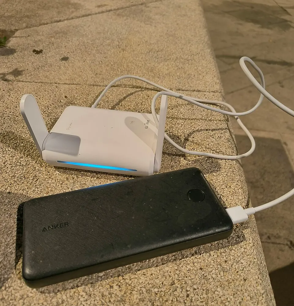 A GL.iNet travel router with a glowing blue status light and an Anker power bank, connected together on a stone ledge outdoors
